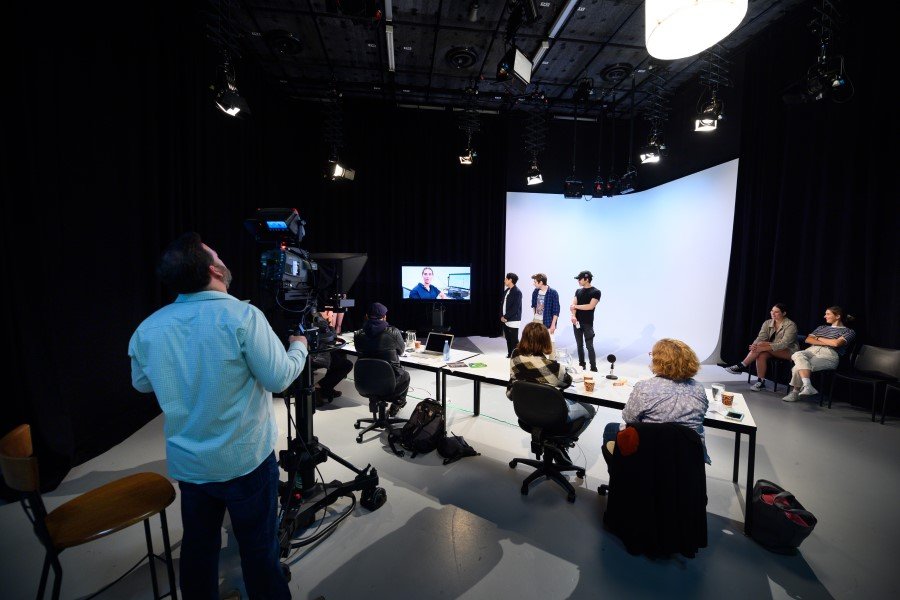 在一個設有大型白色無縫背景牆（Cyclorama）的專業媒體電視廣播演播室中，多名傳理系學生正進行實務操作課程。左側前景有一名攝影師正操作著大型廣播級攝影機，中景的長桌前坐著多名操作控制設備與紀錄的學生，遠處則有三名學生在燈光與白色背景前進行拍攝排練，上方天花板掛滿了專業舞台照明燈具。