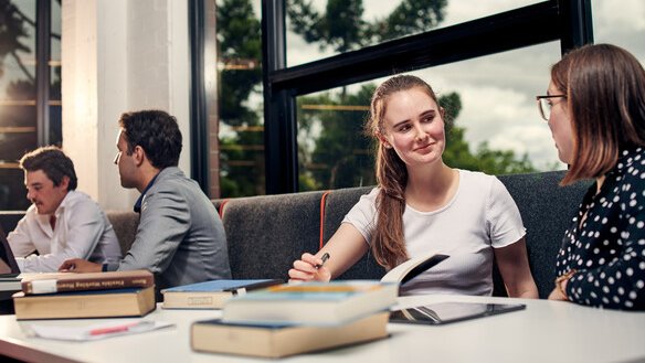 Deakin University商學院的女學生坐在現代化的校園休息區，與同學圍繞著放有課本和筆記本的桌子進行小組討論，背景有寬敞的落地窗。