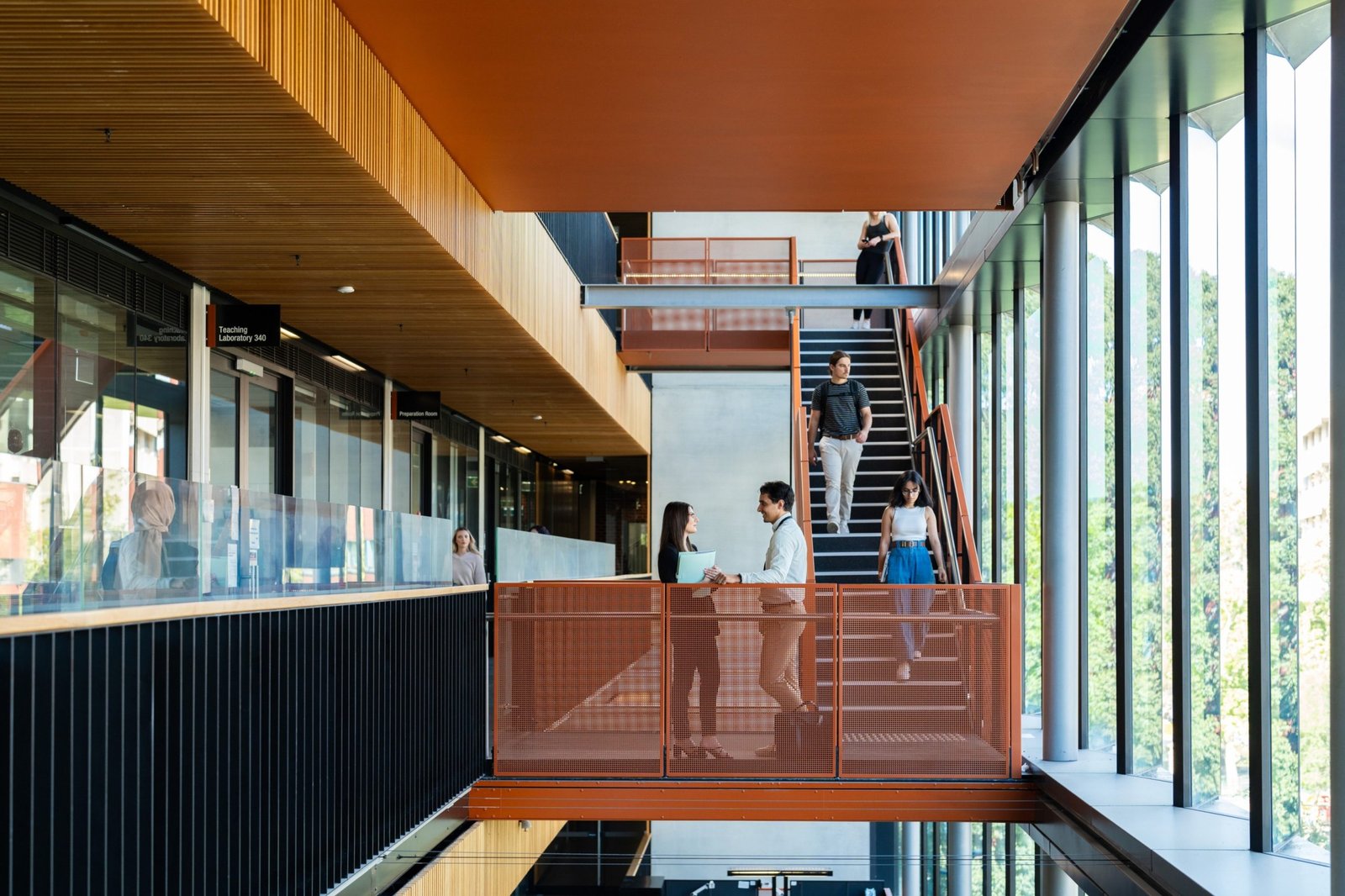 一棟現代化大學校園建築的明亮室內空間。畫面中央有兩位學生在橘色網狀欄杆的走廊上交談，後方有其他學生正在走下樓梯。左側為帶有木質天花板與玻璃隔間的「教學實驗室（Teaching Laboratory 340）」，右側則是大片落地窗，透出室外的綠意樹木。