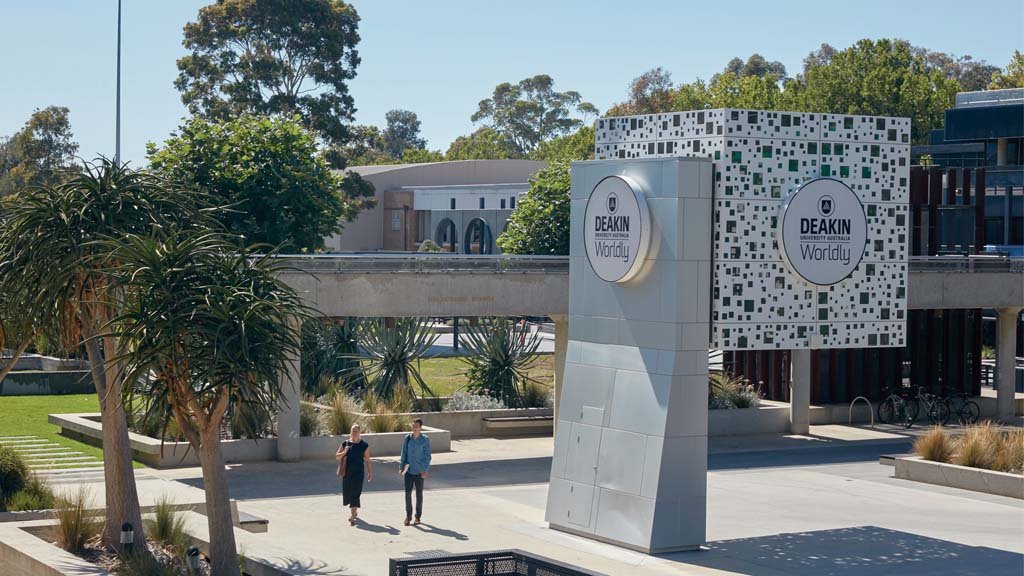 Deakin Univerity校園一景。畫面右側是一座大型的現代白色設計裝置，上方印有圓形的迪肯大學校徽及「Deakin Worldly」字樣，後方相連的立方體外牆佈滿不規則的方形鏤空孔洞。兩名行人（一男一女）正走在前景寬敞的戶外步道上。校園內種植了許多綠色植物與樹木，背景隱約可見一座帶有大型圓拱窗戶的現代教學樓，上方為晴朗的藍天。